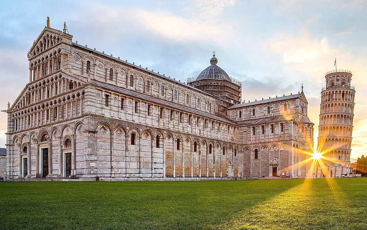 La Cattedrale di Pisa all'alba: la lunga facciata di marmo con il sole che si apre a stella dietro la Torre pendente a destra