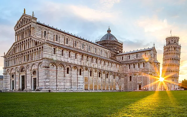 La Cattedrale di Pisa all'alba: la lunga facciata di marmo con il sole che si apre a stella dietro la Torre pendente a destra